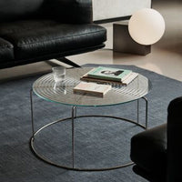 Round glass coffee table with a textured surface, silver metal frame, and placed on a dark blue rug. Modern living room decor.
