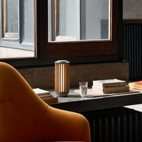 Warm orange armchair, modern wooden lamp with vertical stripes, neutral-toned table with books and a glass.