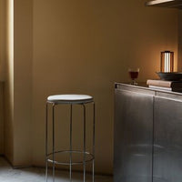 Minimalist chrome bar stool with a white seat against a warm, muted yellow wall and subtle gray flooring.