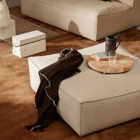 Beige ottoman with black accent scarf on brown rug, complemented by wooden serving tray and clear glass on the table.