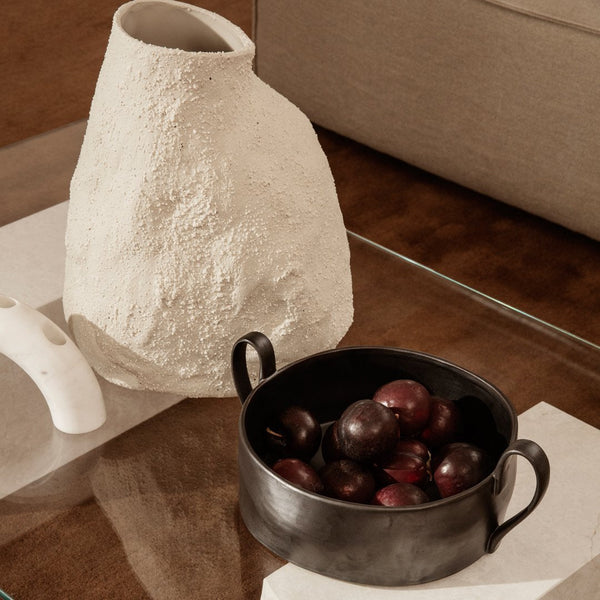 Textured white vase beside a black bowl filled with deep purple fruits, situated on a clear glass table.