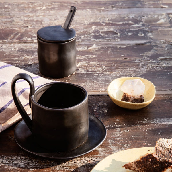 Black ceramic cup and saucer set with a matte finish, paired with a textured black sugar jar and a pastel yellow dish.