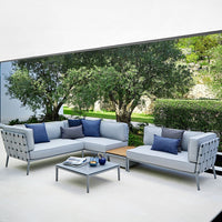 Gray outdoor sectional sofa with blue and purple accent pillows, set against a lush green backdrop.