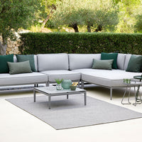 Gray outdoor sectional sofa with green accent pillows, placed on a light gray rug in a lush garden setting.