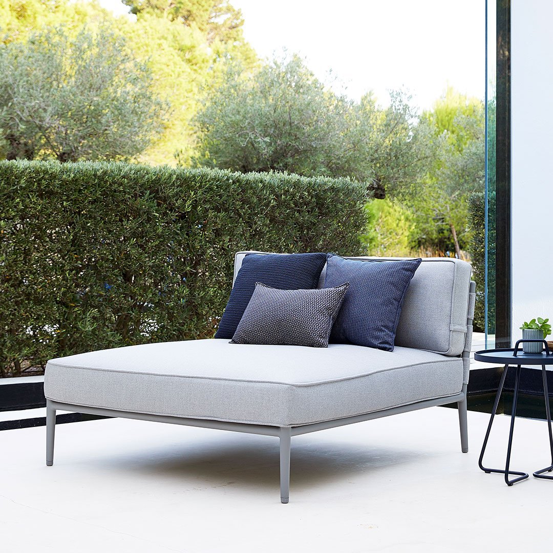 Neutral grey chaise lounge with dark blue and light grey accent pillows, set in a lush green outdoor space.