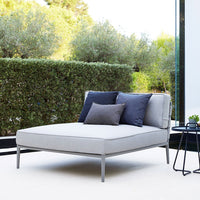 Neutral grey chaise lounge with dark blue and light grey accent pillows, set in a lush green outdoor space.