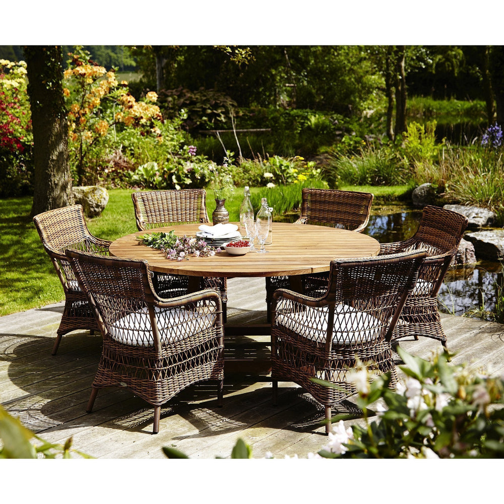 Round outdoor dining set featuring brown wicker chairs with striped cushions against a lush green garden backdrop.