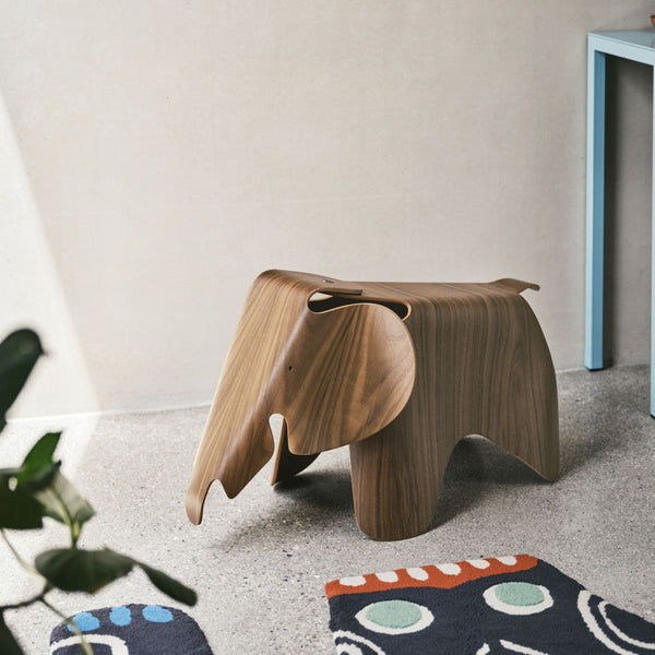 Wooden elephant stool in warm brown tones, featuring smooth curves and minimalistic design, set against colorful patterned rugs.