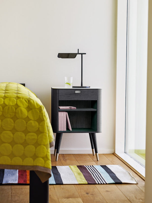 Modern black nightstand with sleek lines beside a yellow-patterned quilt and striped multi-color rug.