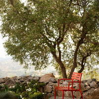 Bright red metal chair with a simple design, surrounded by lush green foliage and a natural stone background.