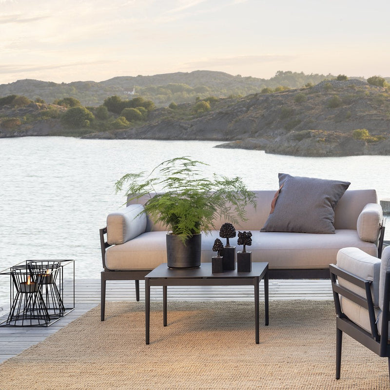 Neutral-toned outdoor sofa with gray cushions and black metal accents, set against a serene lakeside backdrop.