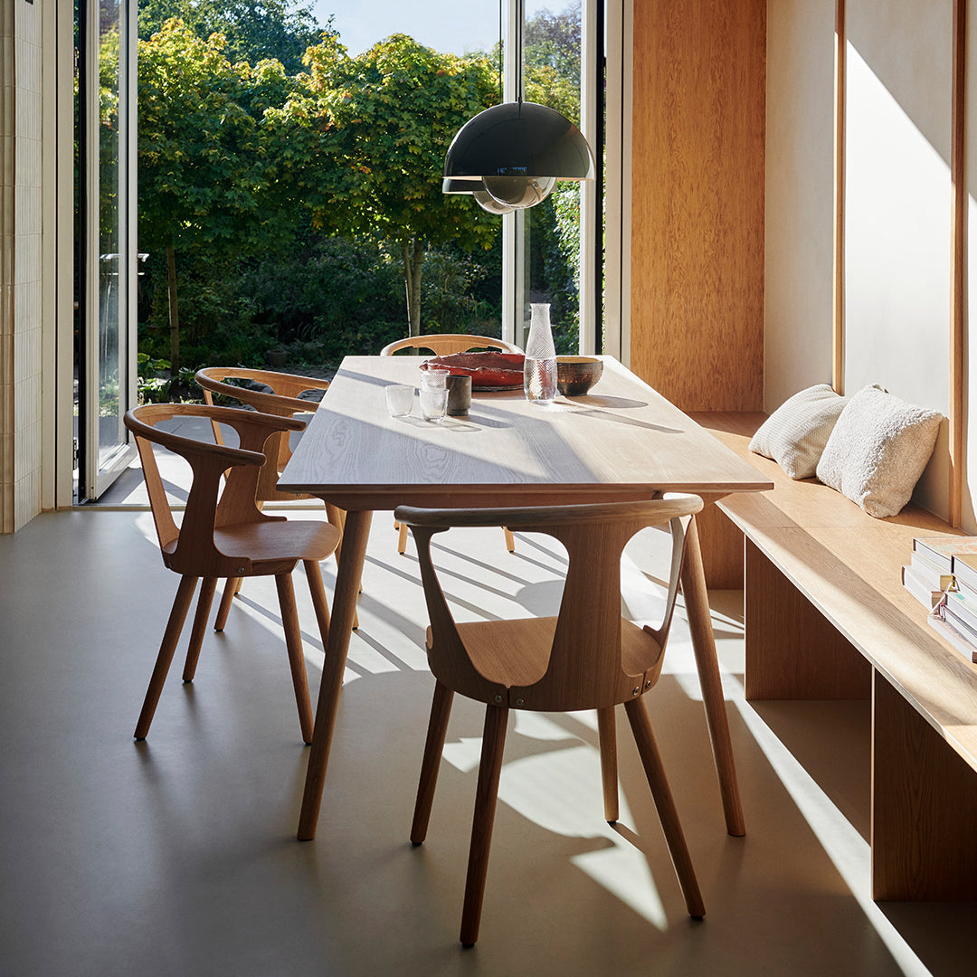 Modern wooden dining table with natural finish, accompanied by light wood chairs, set in a bright, green-accented space.