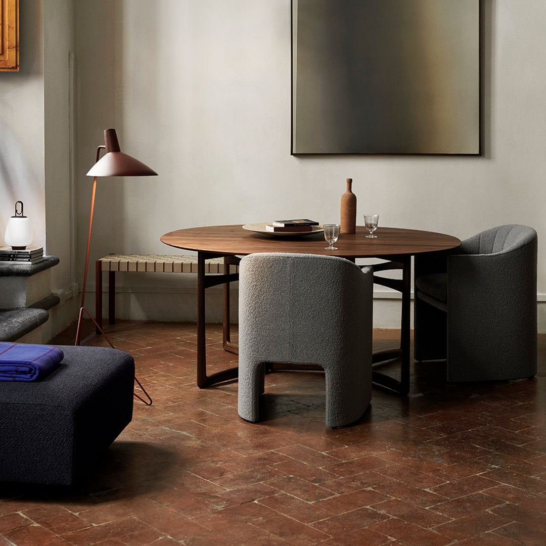 Round wooden dining table with gray upholstered chairs, featuring earthy tones and a textured floor pattern.