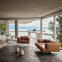 Modern brown leather sofa in a stylish room with natural stone flooring and large glass windows showcasing a serene lake view.