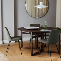 Round brown dining table with green upholstered chairs, set against a neutral wall and a patterned rug.