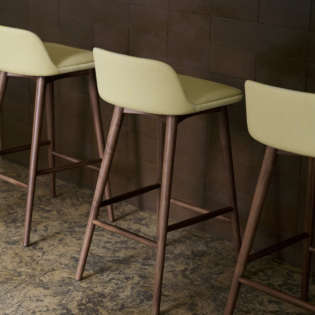 Light green upholstered bar stools with wooden legs against a textured brown wall and patterned floor.