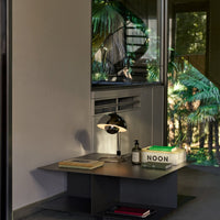 Modern black table with sleek design, accented by a white lamp and green book, surrounded by lush greenery.
