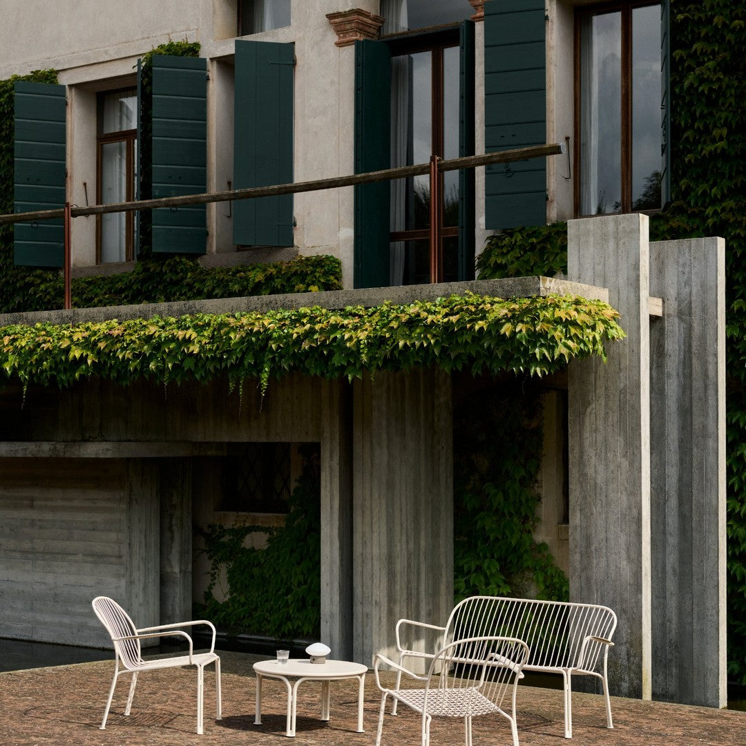 White metal patio furniture with a minimalist design, set against a backdrop of green foliage and rustic gray concrete.