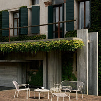 White metal patio furniture with a minimalist design, set against a backdrop of green foliage and rustic gray concrete.