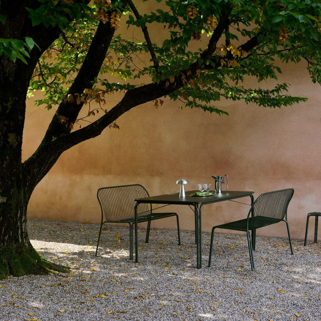 Modern green outdoor dining set against a warm, muted pink background with textured gravel and lush greenery.