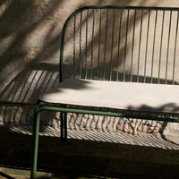 Green metal bench with a light beige cushion, featuring sleek vertical lines and subtle shadows.