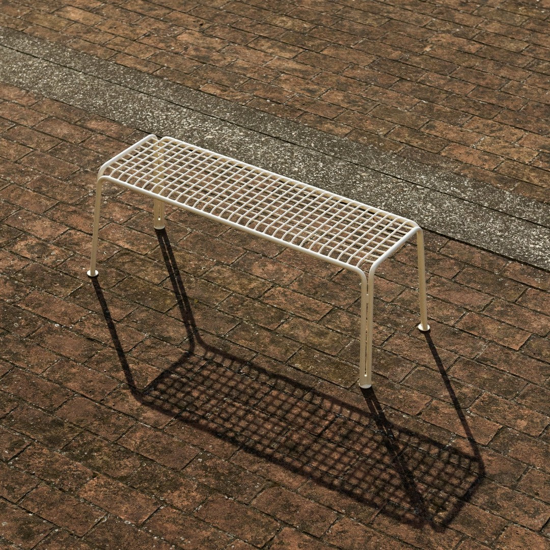 Beige metal bench with a grid pattern, displayed on a textured brown outdoor surface.