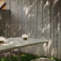 Beige table with round light fixtures, green grass, and textured gray wood wall in soft, natural light.