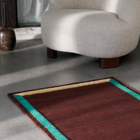 Brown rug with turquoise and yellow borders, placed beside a light-colored chair on a concrete floor.