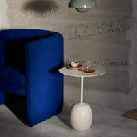 Blue upholstered chair with a white round side table displaying a glass and wooden coaster against a textured gray background.