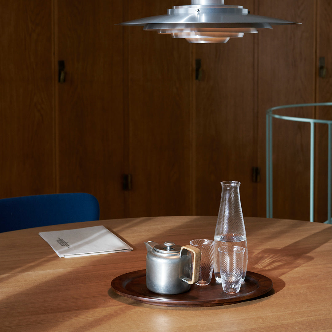 Silver teapot and glass carafe on a brown wooden tray, set against a warm, minimalist background.