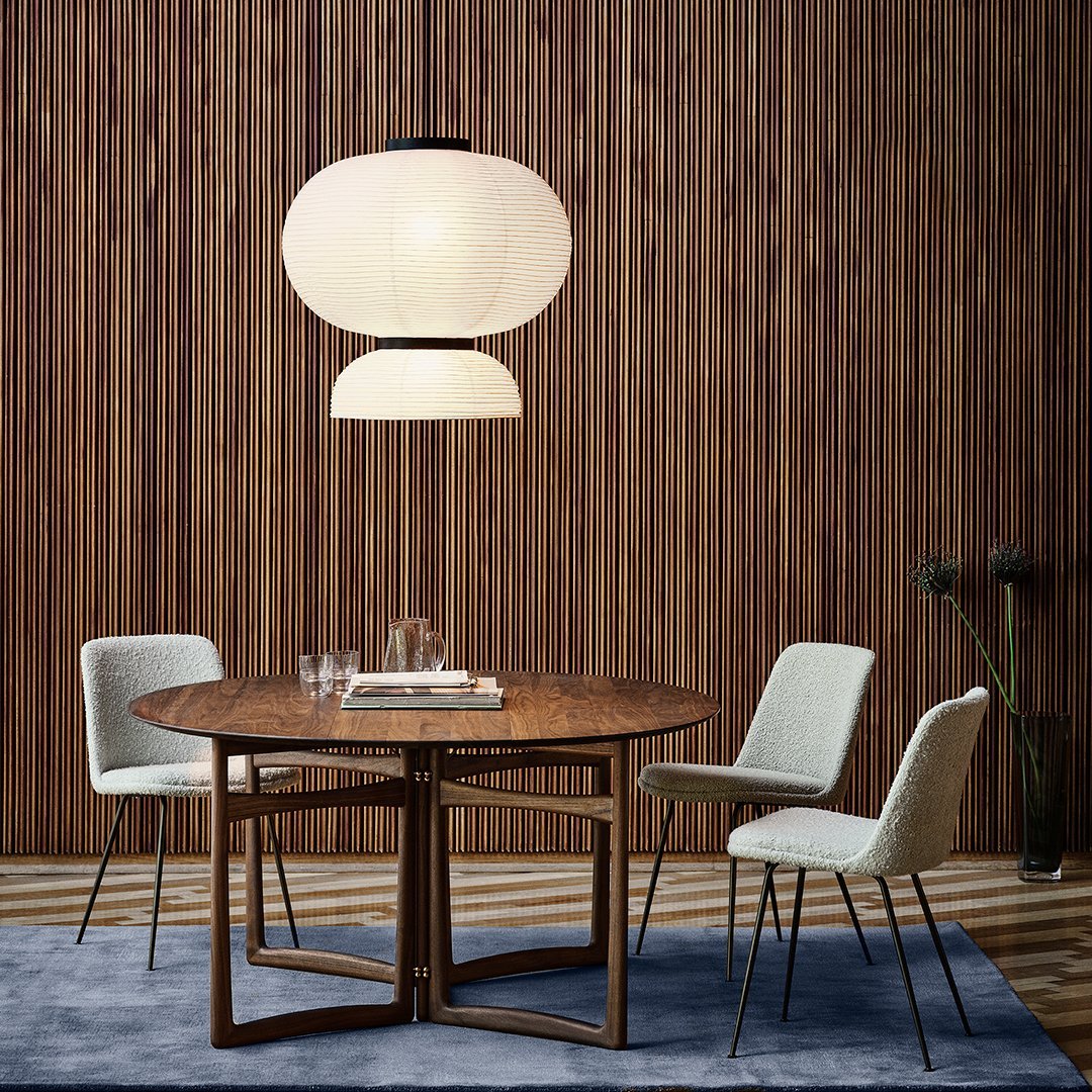Round wooden dining table with beige chairs, set against a textured wooden backdrop and a soft blue rug. Stylish pendant light above.