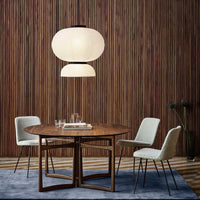 Round wooden dining table with beige chairs, set against a textured wooden backdrop and a soft blue rug. Stylish pendant light above.