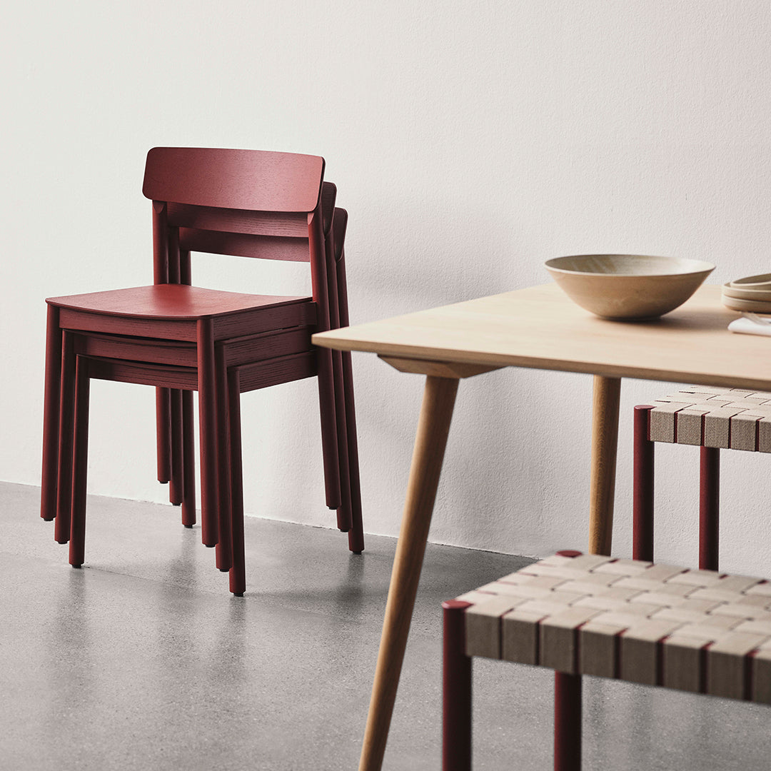 Rustic red stacked chairs next to a light wood table with woven seat detailing, set against a neutral background.