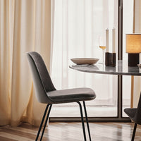 Stylish grey velvet chair with sleek black legs, set against soft beige curtains and a minimalistic marble table.