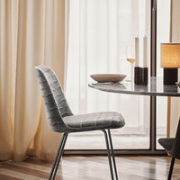 Gray quilted dining chair standing next to a minimalist round table, with soft beige curtains in the background.