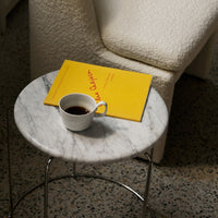 White marble table with a yellow book, dark coffee, and a light gray cup on a speckled floor. Modern, elegant colors.