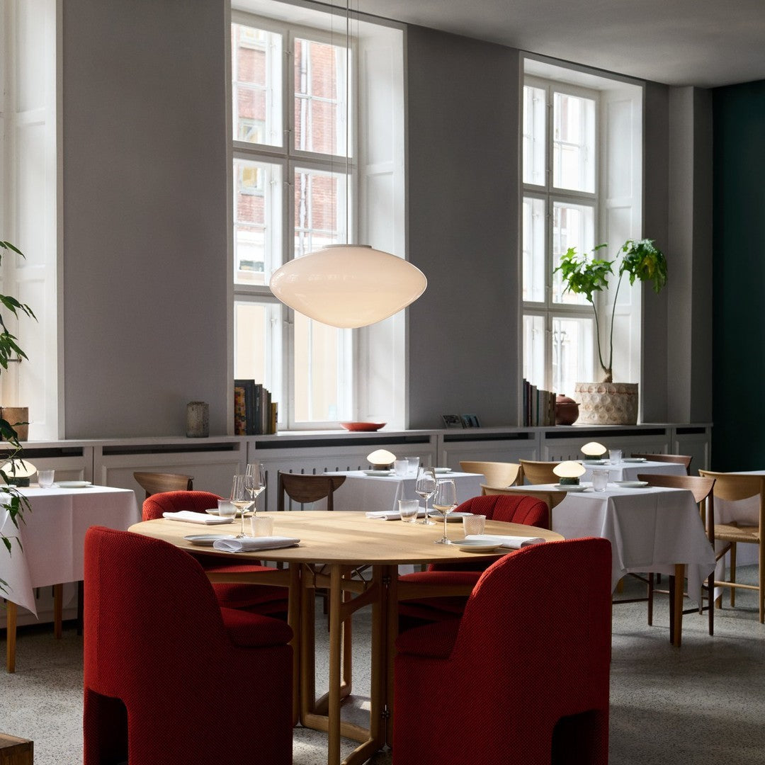 Red upholstered chairs around a round wooden table in a light-filled, minimalistic restaurant interior with white linens.