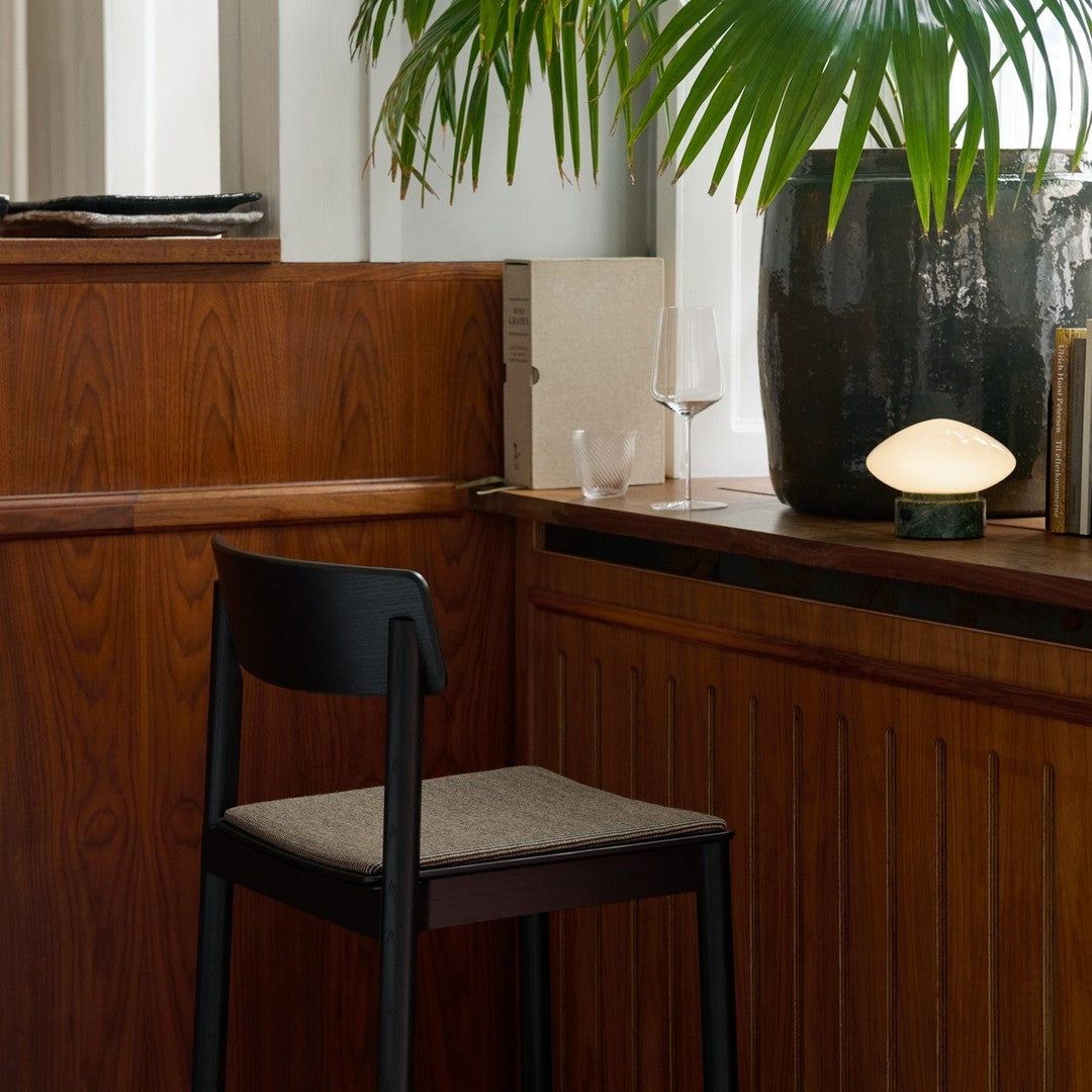 Black wooden chair with a beige fabric seat against rich brown wood paneling, accented by green foliage and soft lighting.