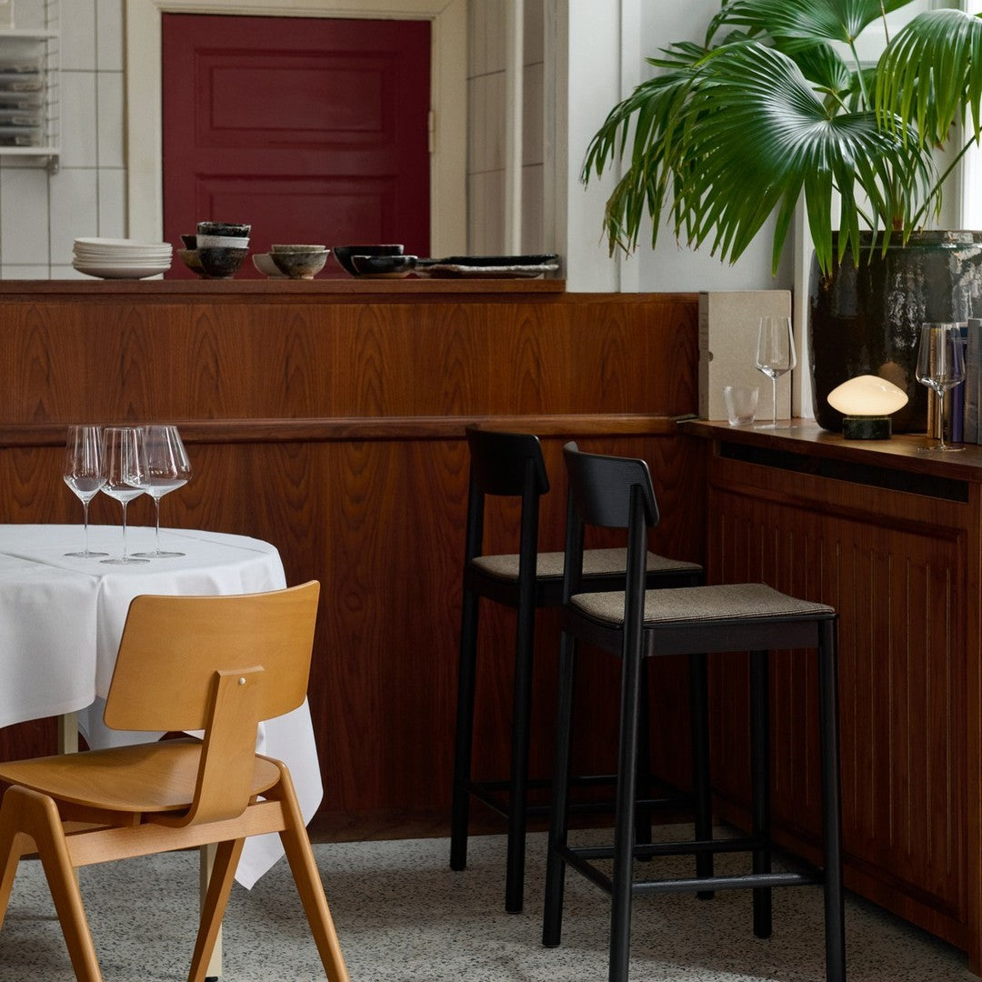 Modern dining scene featuring warm wood tones, a crisp white tablecloth, black chairs, and green plant accents.