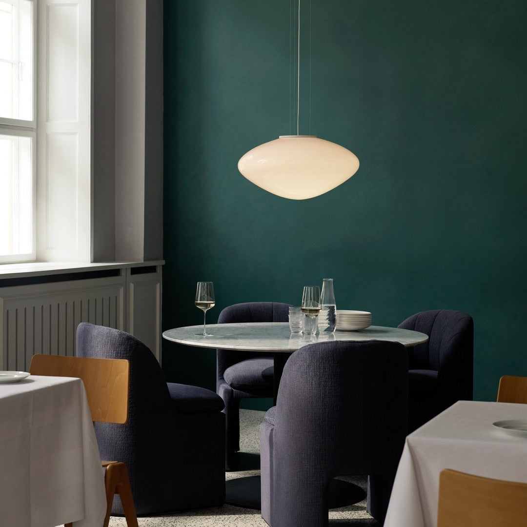Sleek white pendant lamp hanging above a round marble table, surrounded by deep navy chairs against a rich green wall.