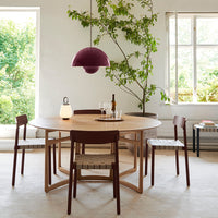 "Round wooden table with burgundy pendant light, natural wood chairs, and greenery backdrop. Modern and inviting design."