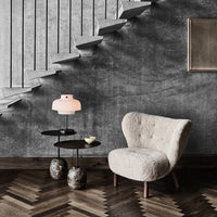 Cozy chair with a textured beige fabric, sleek black side table, and warm pink lamp against a gray wall and wooden herringbone floor.