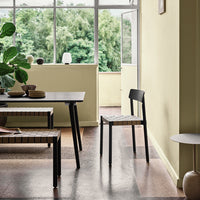 Black chair with woven seat, set against a soft yellow wall and earthy-toned patterned flooring. Bright natural light.