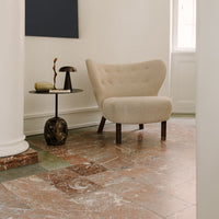 Beige upholstered chair with soft curves, paired with a dark textured side table on a marbled floor with earthy tones.