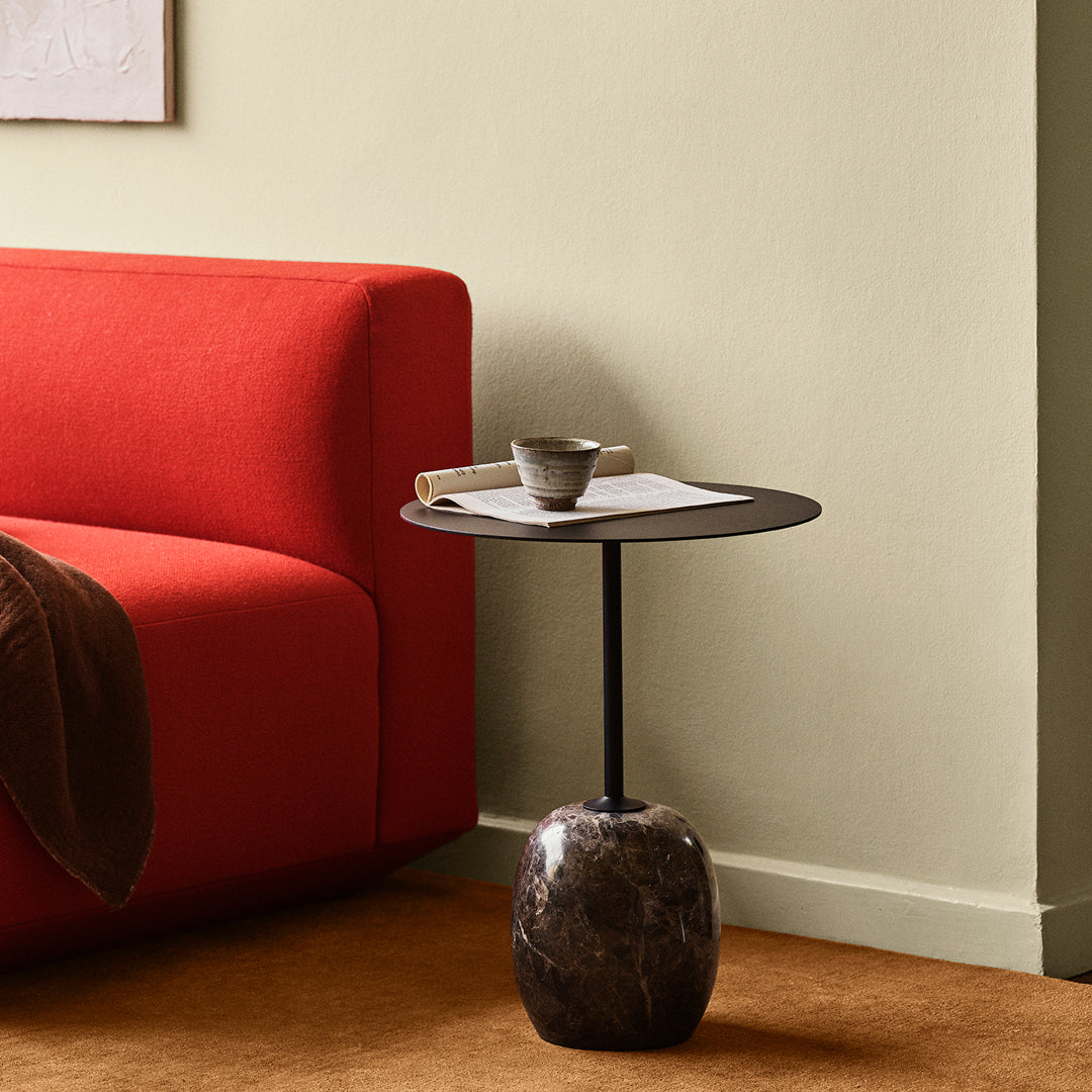 Black circular tabletop on a textured dark stone base, alongside a vibrant red sofa and earthy brown floor.