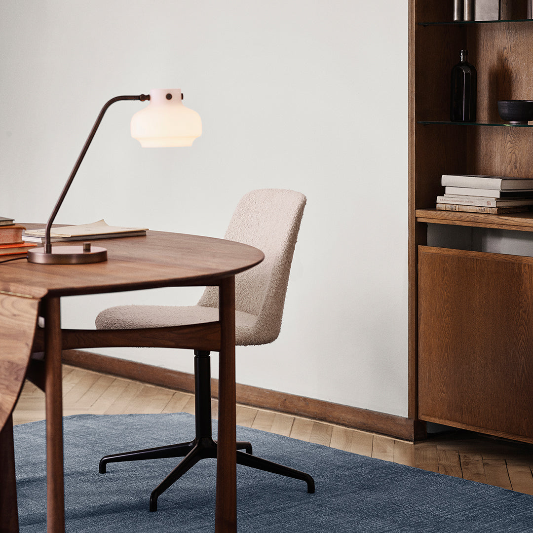 Elegant white lamp with a glass shade, brown wooden desk, neutral fabric chair, and blue textured rug. Modern home office decor.