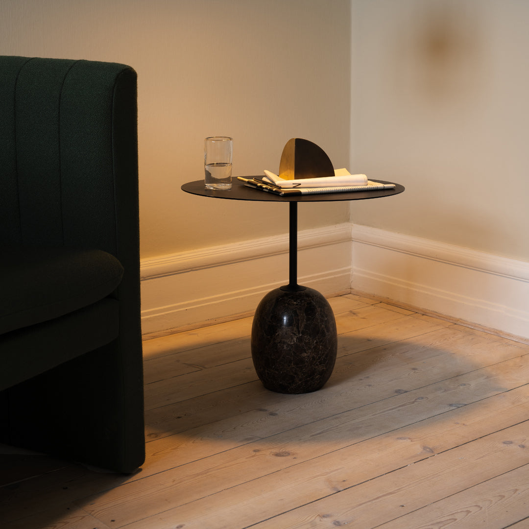 Black round side table with a glossy top and an oval marble base, set against a light wooden floor and green upholstery.