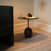 Black round side table with a glossy top and an oval marble base, set against a light wooden floor and green upholstery.