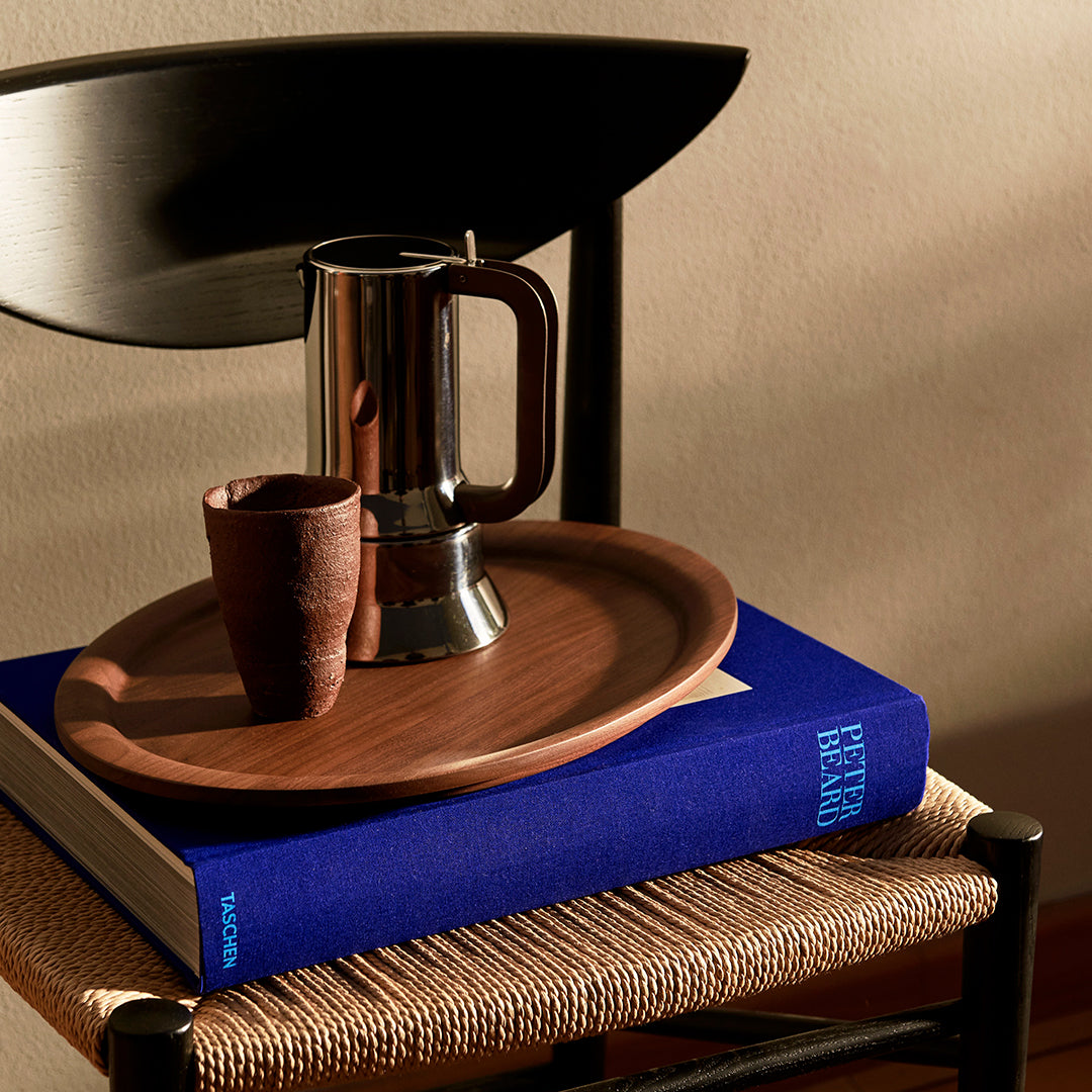 Wooden tray in warm brown with a silver coffee pot and dark clay cup on a blue book, creating a cozy, stylish setup.
