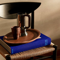 Wooden tray in warm brown with a silver coffee pot and dark clay cup on a blue book, creating a cozy, stylish setup.